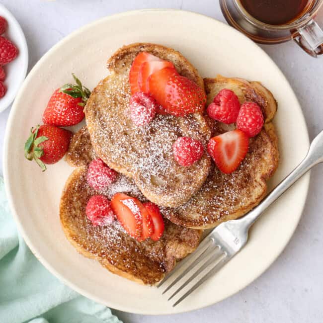 Vegan French Toast on a plate topped with strawberries, with more toppings and another plate of the french toast surrounding.