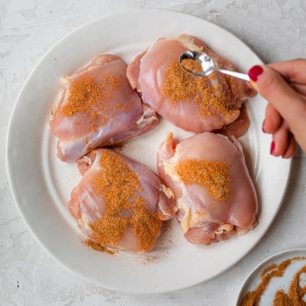 A spoon full of the seasoning used to make the chicken thighs being sprinkled across the plate of four chicken thighs,