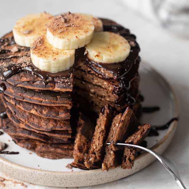 Chocolate protein pancakes stacked on a plate with banana slices on top and a fork taking a bite.
