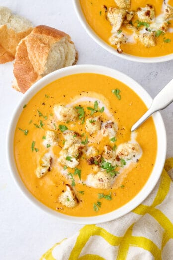 Two bowls of soup with a little extra swirl of coconut milk and some roasted cauliflower spooned on top, garnished with cilantro, crust bread nearby.