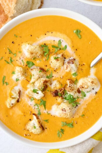 Two bowls of soup with a little extra swirl of coconut milk and some roasted cauliflower spooned on top, garnished with cilantro, crust bread nearby.