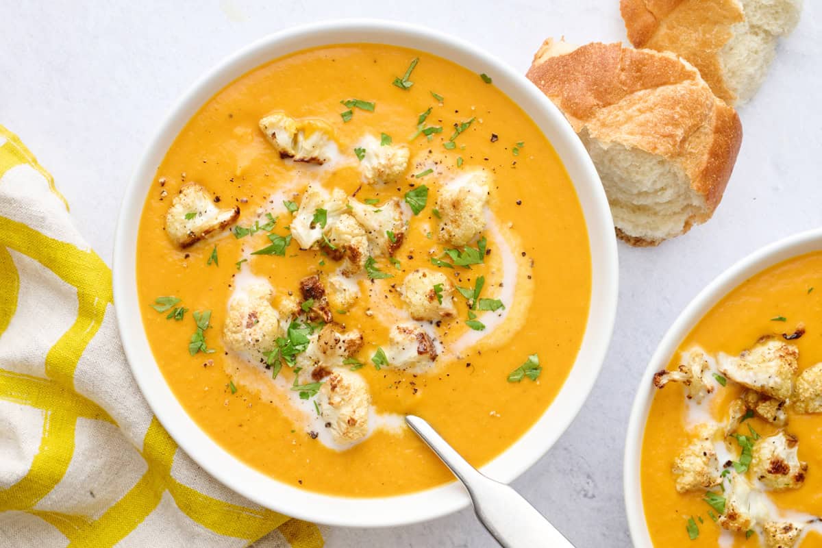 Two bowls of soup with a little extra swirl of coconut milk and some roasted cauliflower spooned on top, garnished with cilantro, crust bread nearby.