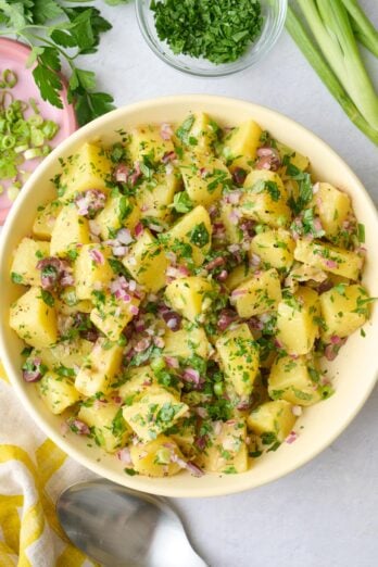 Potato salad in a serving bowl with parsley on top and extra ingredients around.