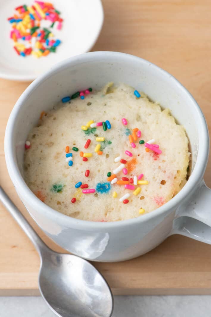Vanilla mug cake with extra sprinkles on top and a spoon nearby.