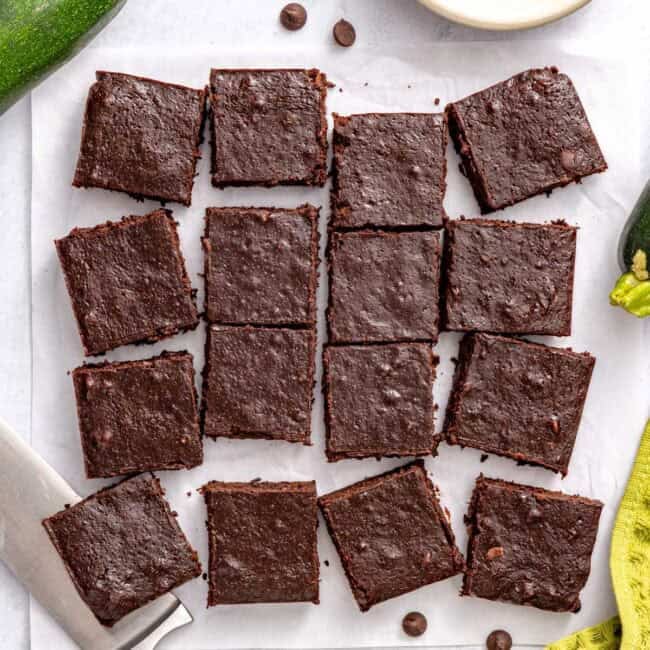 Zucchini brownies on parchment paper cut into 16 squares with a few pulled away with a zucchini and chocolate chips nearby.