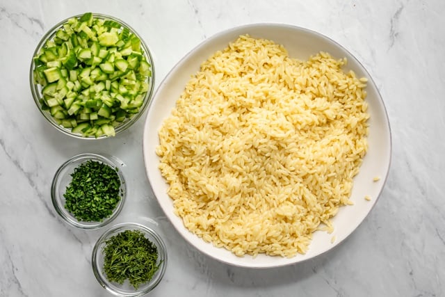 Orzo salad tossed in the dressing in a large bowl with cucumbers and herbs on the side