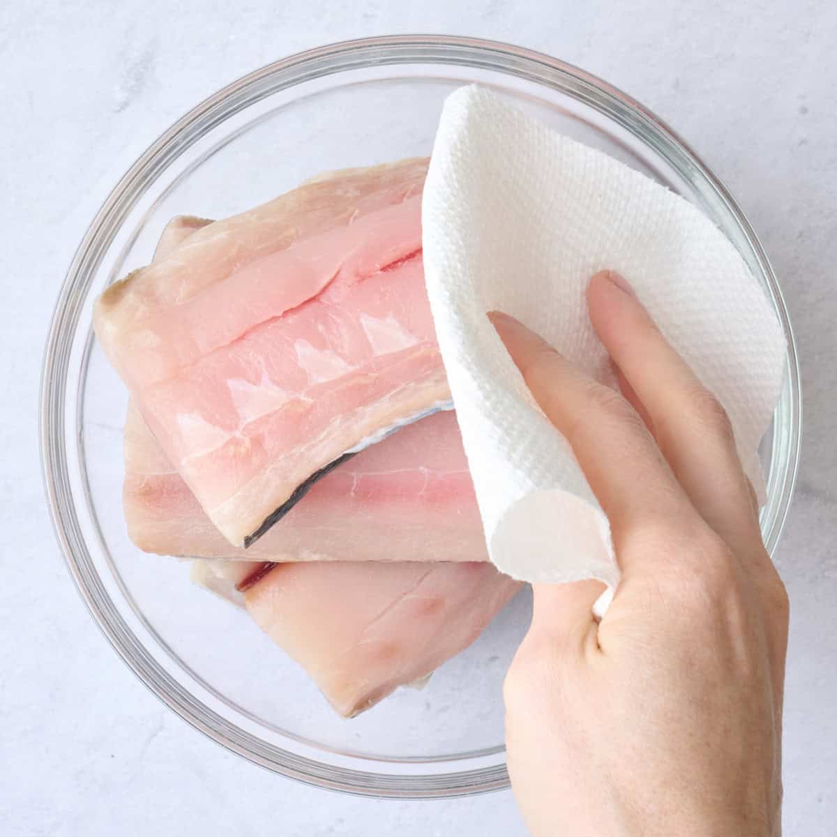 Fish fillets in a bowl being pat dry with a paper towel.