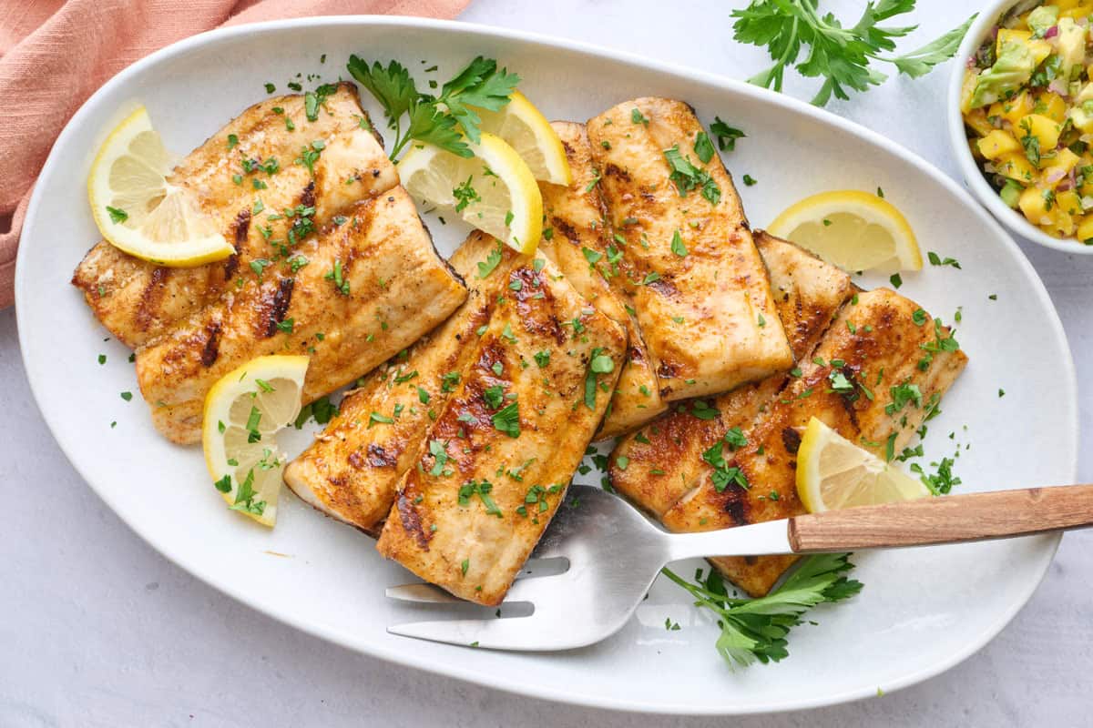 Grilled mahi mahi fillets on a large platter garnished with chopped parsley and lemon wedges, small bowl of mango salsa nearby.