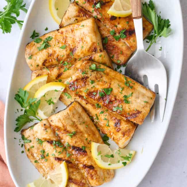 Grilled mahi mahi fillets on a large platter garnished with chopped parsley and lemon wedges, small bowl of mango salsa nearby.