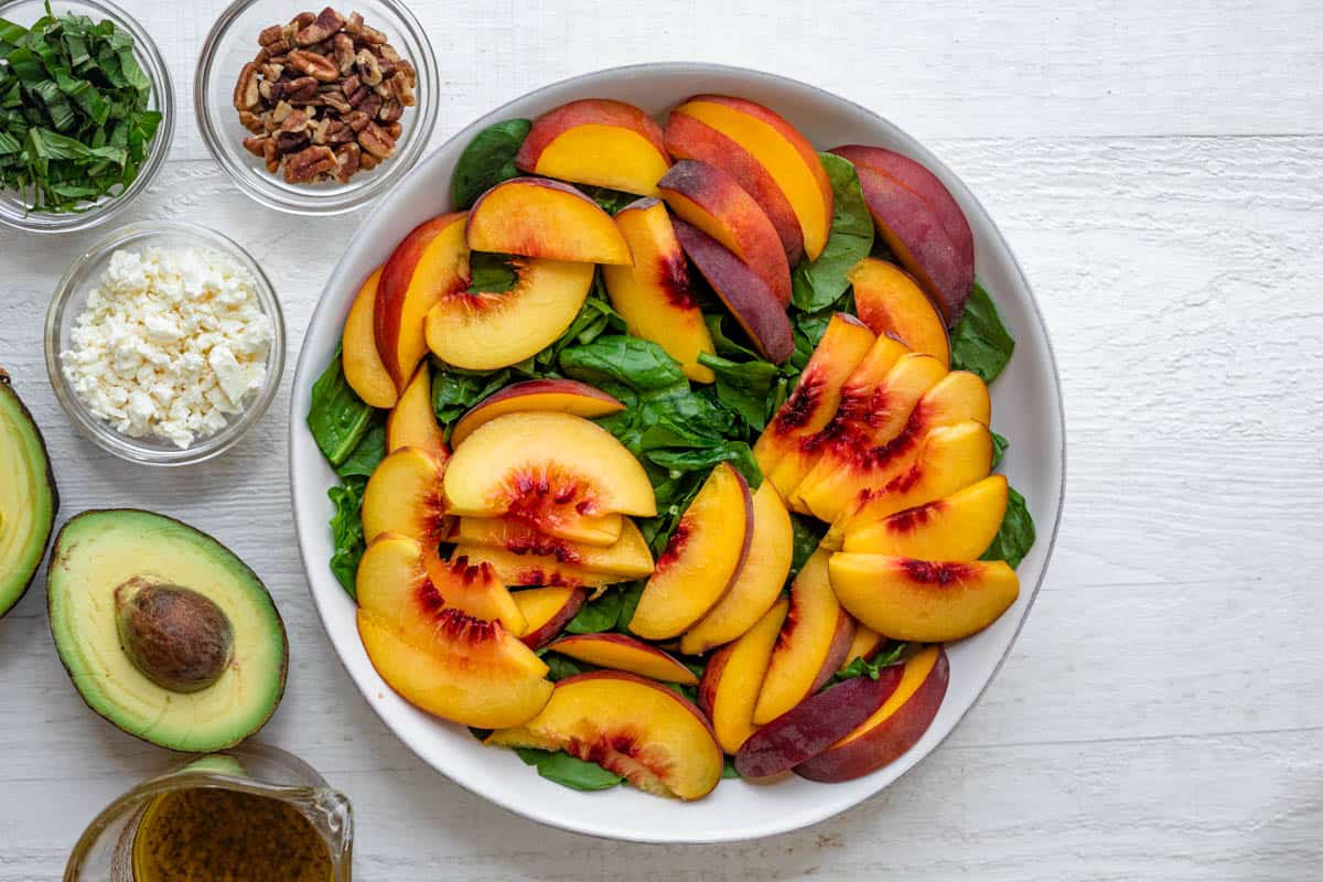 Large bowl of spinach and peaches with avocados, basil, pecans, feta cheese and dressing on the side