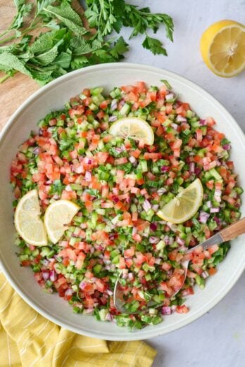 Shirazi salad in a serving bowl with a serving spoon dipped inside, fresh lemon and extra herbs nearby.