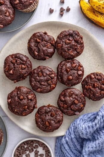 Chocolate banana muffins on a plate with a few on small plates nearby, bananas and small dish of chocolate chips around.