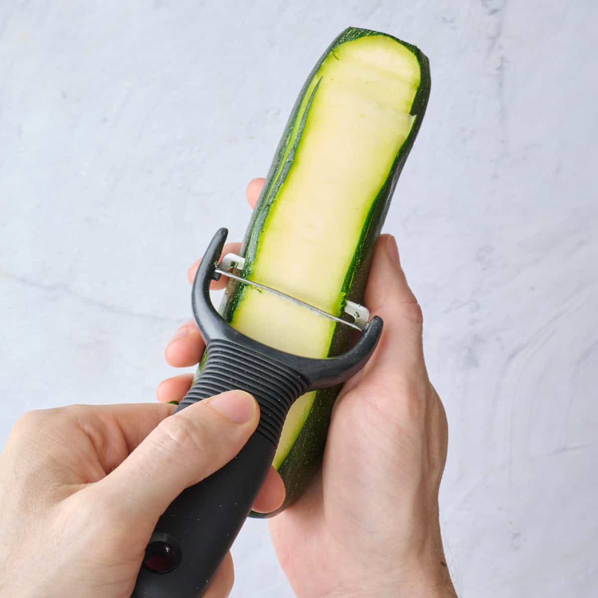 Hand holding a partially sliced zucchini with a Y-Peeler slicing off a piece.