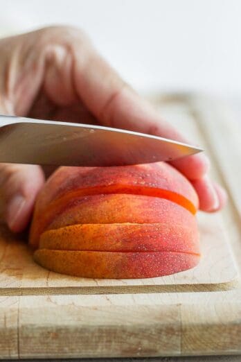 Half of a peach face down on a cutting board with a knife cutting slices.
