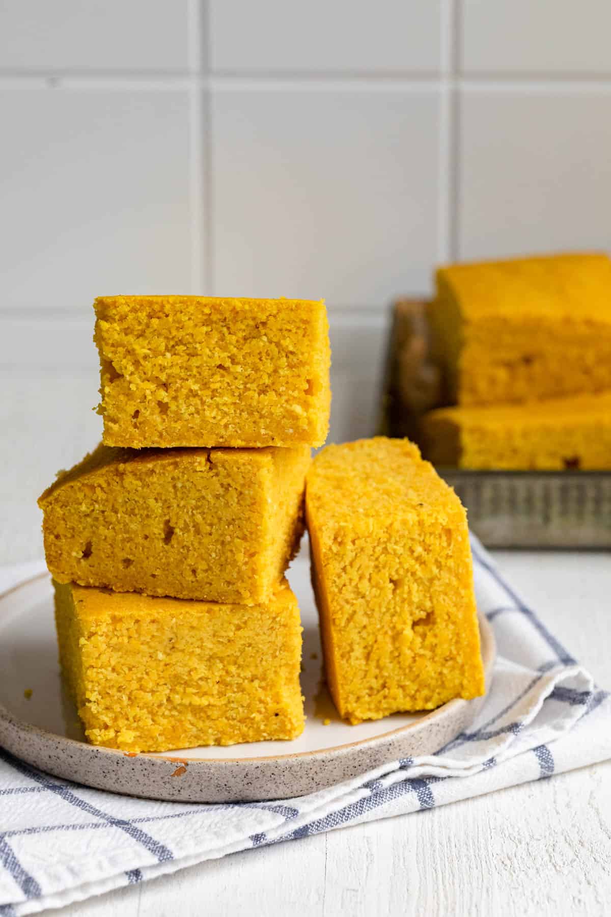 Vegan cornbread stacked on a plate with tray of cornbread in background