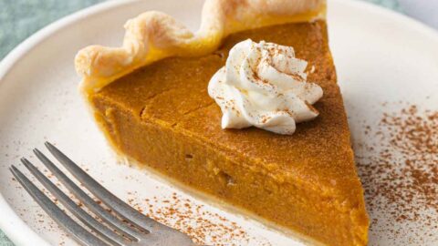 Slice of pumpkin pie on a small plate with whipped cream on top with cinnamon sprinkled on top and a fork sitting on plate.
