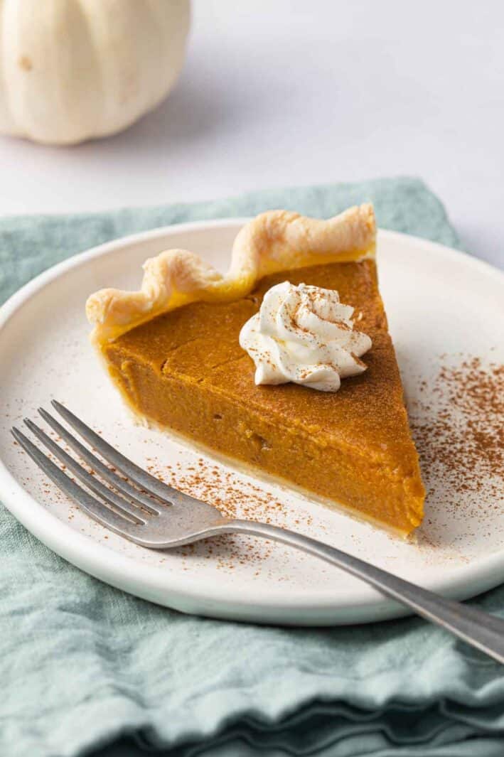 Slice of pumpkin pie on a small plate with whipped cream on top with cinnamon sprinkled on top and a fork sitting on plate.