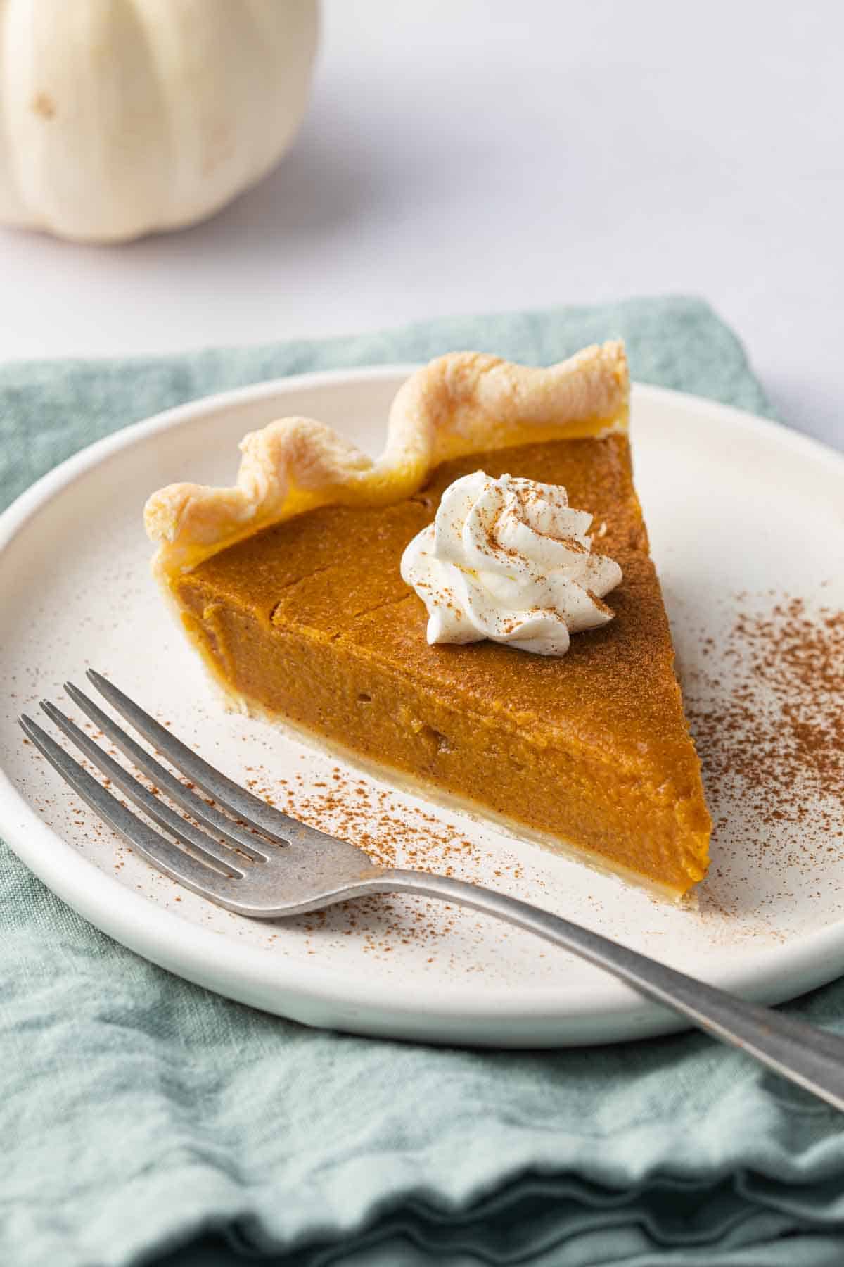 Slice of pumpkin pie on a small plate with whipped cream on top with cinnamon sprinkled on top and a fork sitting on plate.