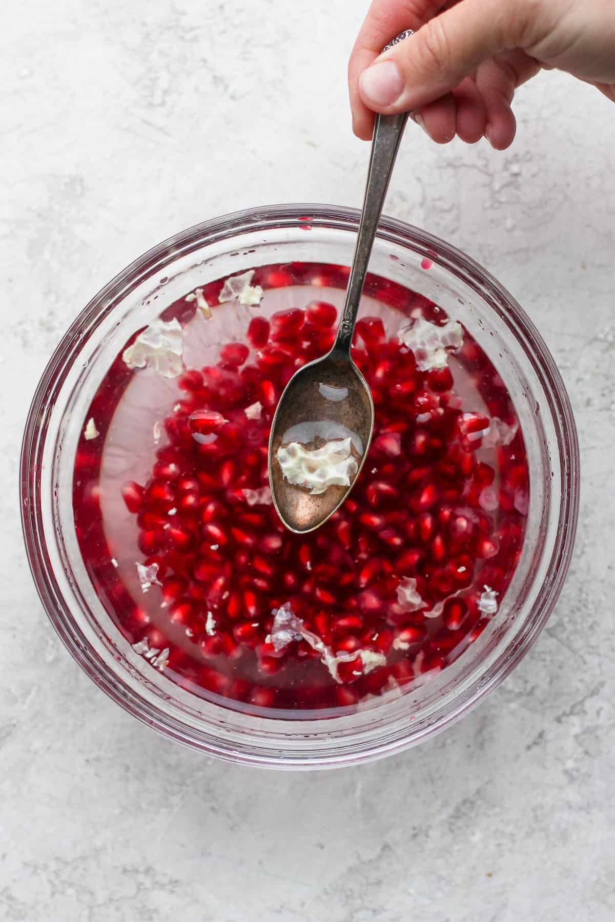 Pomegranate arils in a large bowl with the white pith floating to top and spoon removing them