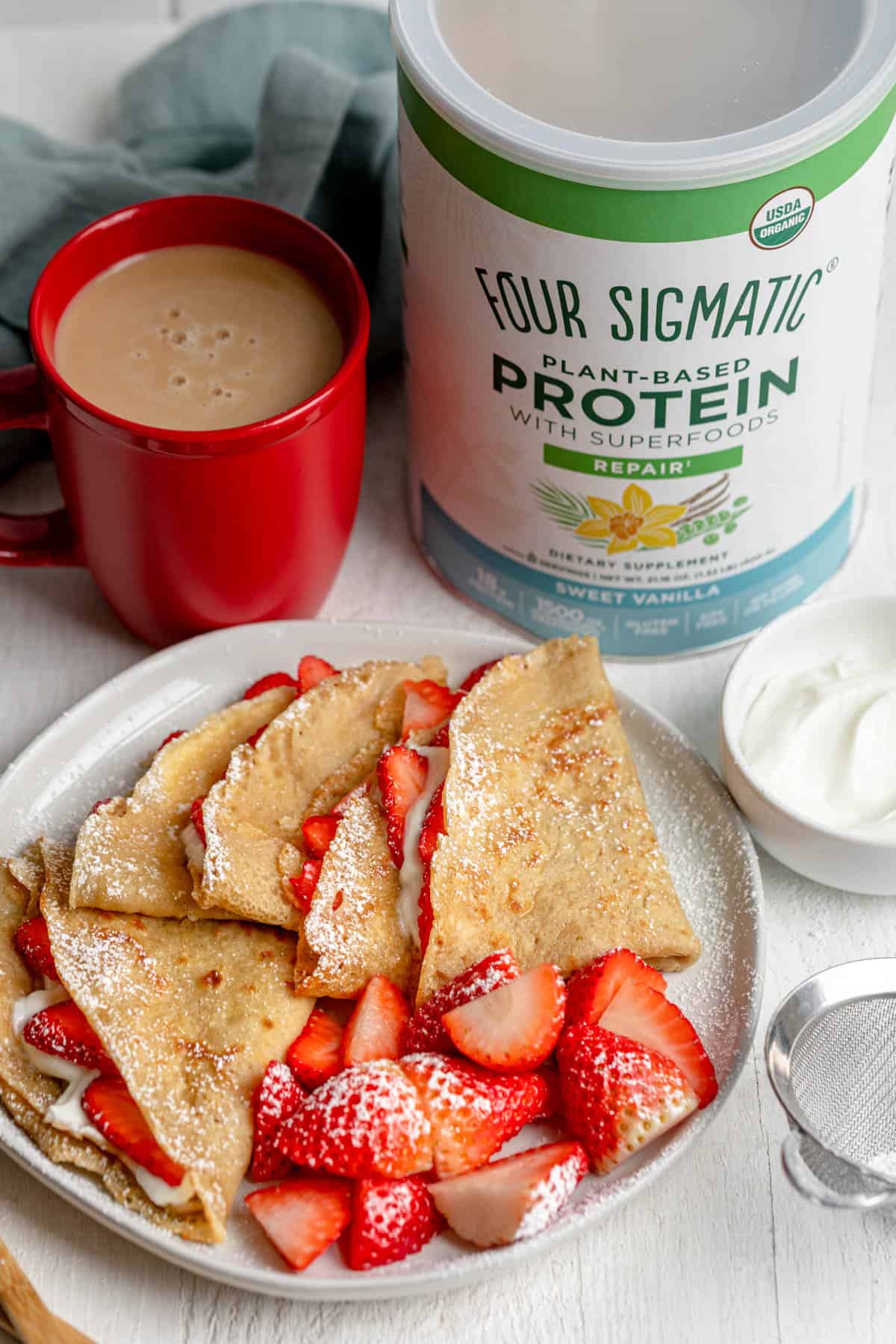 Protein crepes on a plate with strawberries and cream, red coffee mug in background and Four Sigmatic protein powder in background