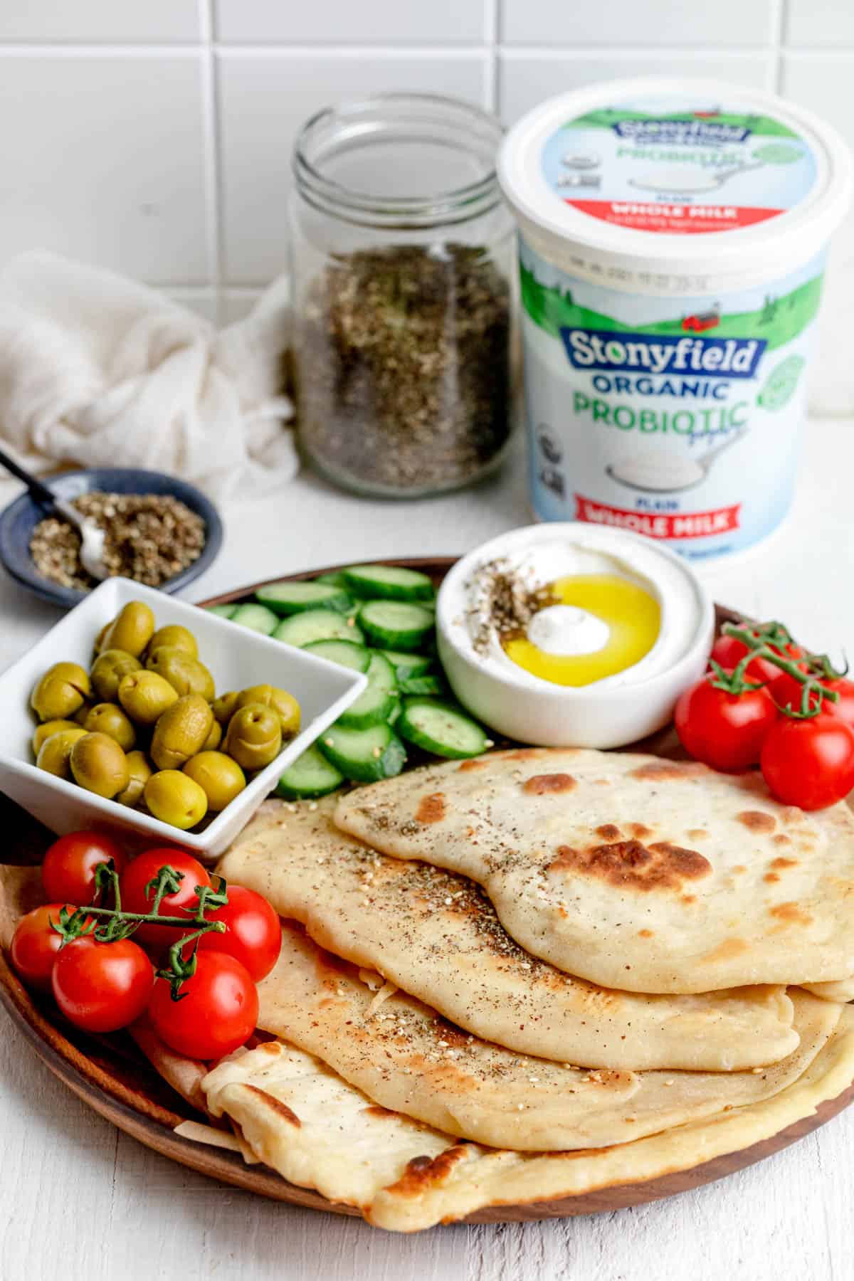 Wooden tray of yogurt flatbread with vegetables and labneh dip, with Stonyfield yogurt in background and zaatar