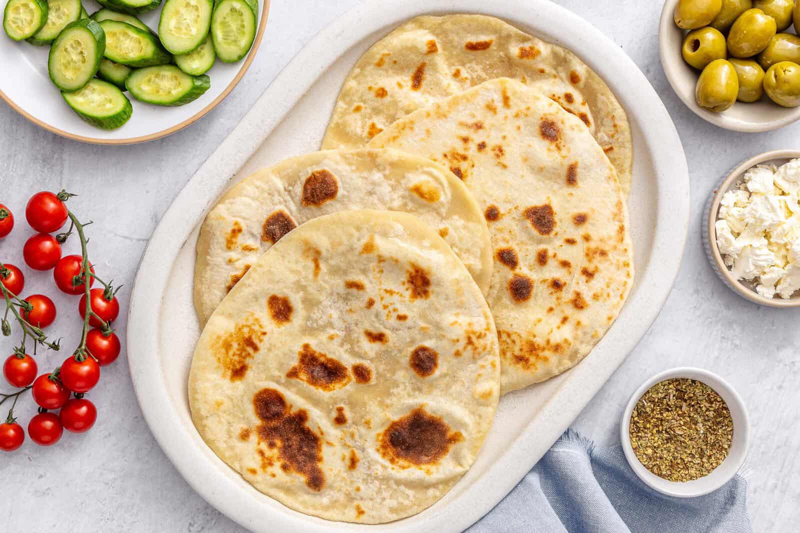 Yogurt flatbreads on a narrow platter with sides of sliced Persian cucumbers, green olives, tomatoes, bowl of crumbled feta cheese.