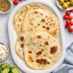 Yogurt flatbreads on a narrow platter with sides of sliced Persian cucumbers, green olives, tomatoes, bowl of crumbled feta cheese.