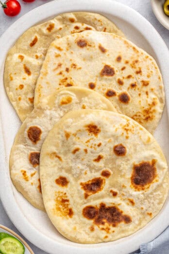 Yogurt flatbreads on a narrow platter with sides of sliced Persian cucumbers, green olives, tomatoes, bowl of crumbled feta cheese.