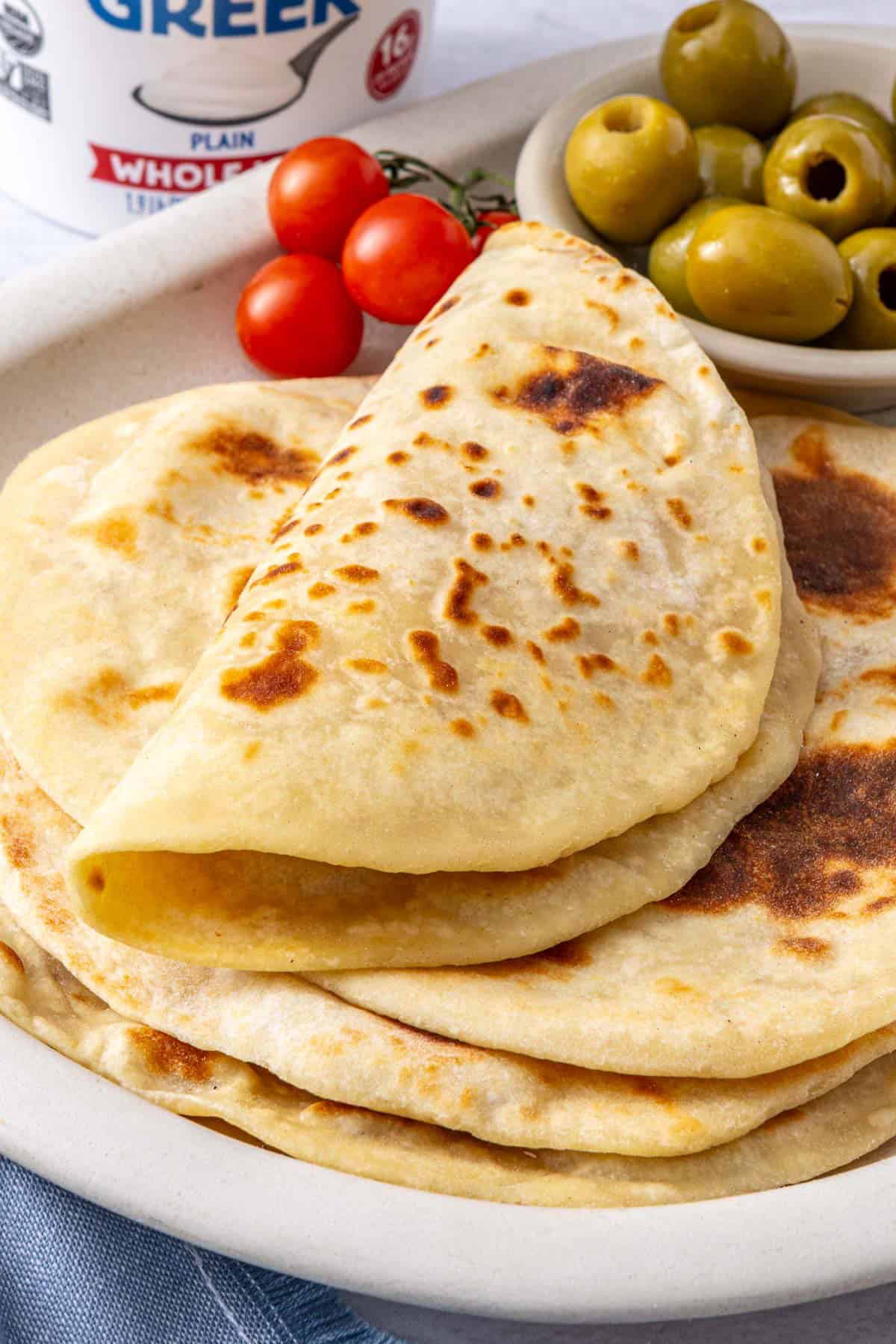 Yogurt flatbreads on a narrow platter, with the top one folded over, with sides of sliced Persian cucumbers, green olives, tomatoes, bowl of crumbled feta cheese.