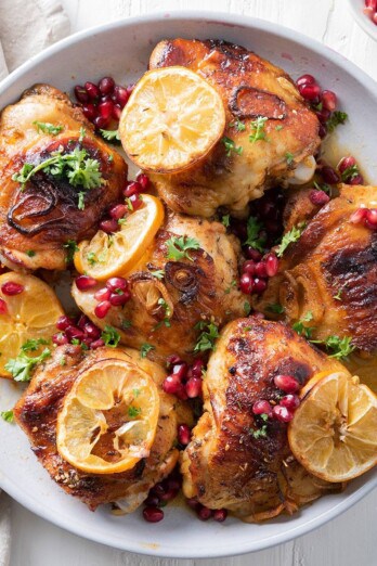 Pomegranate chicken thighs in a serving bowl with shallots, lemons, and garnish.