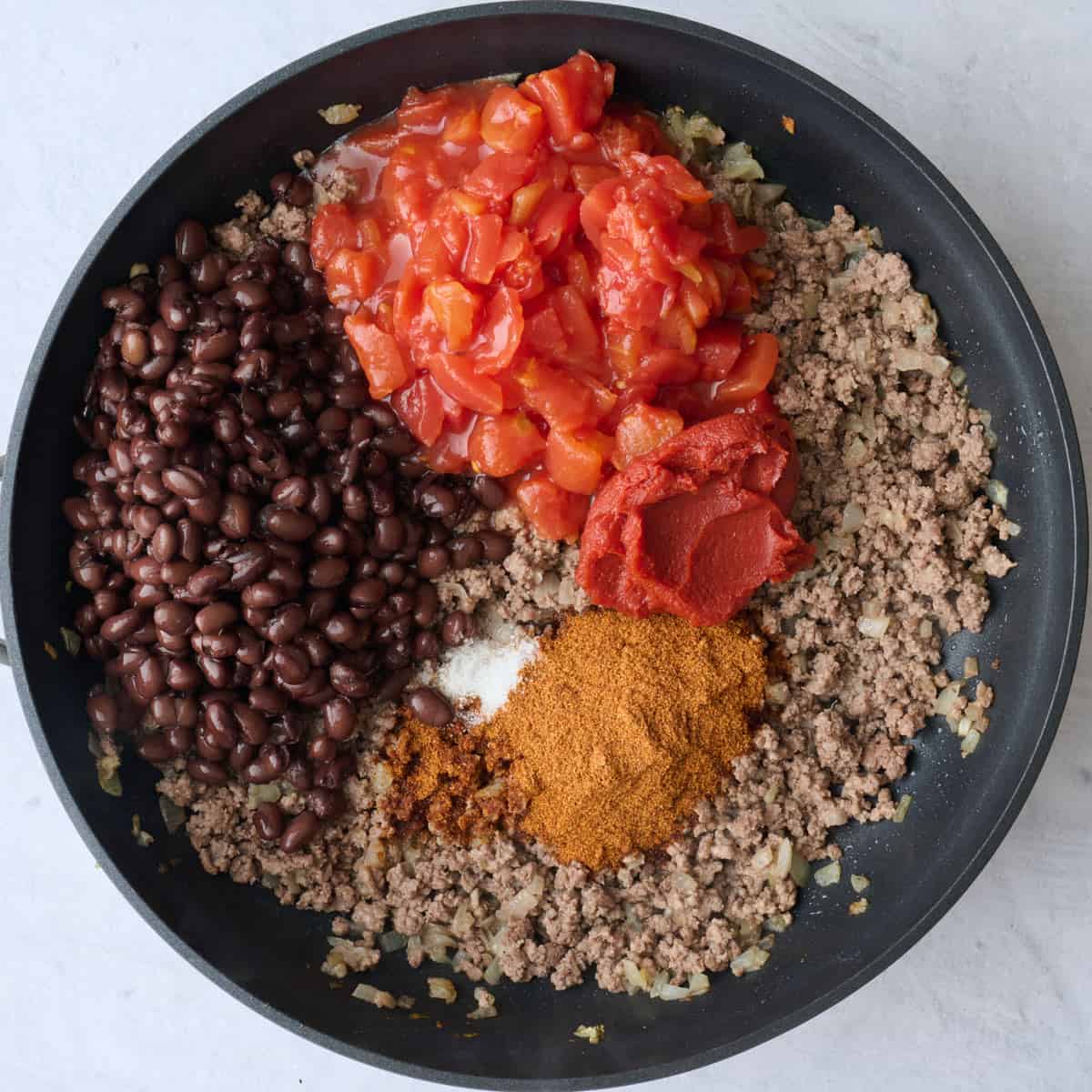 Beef in the skillet after cooking, with taco seasoning, salt, tomato paste, water, beans, and tomatoes added on top.