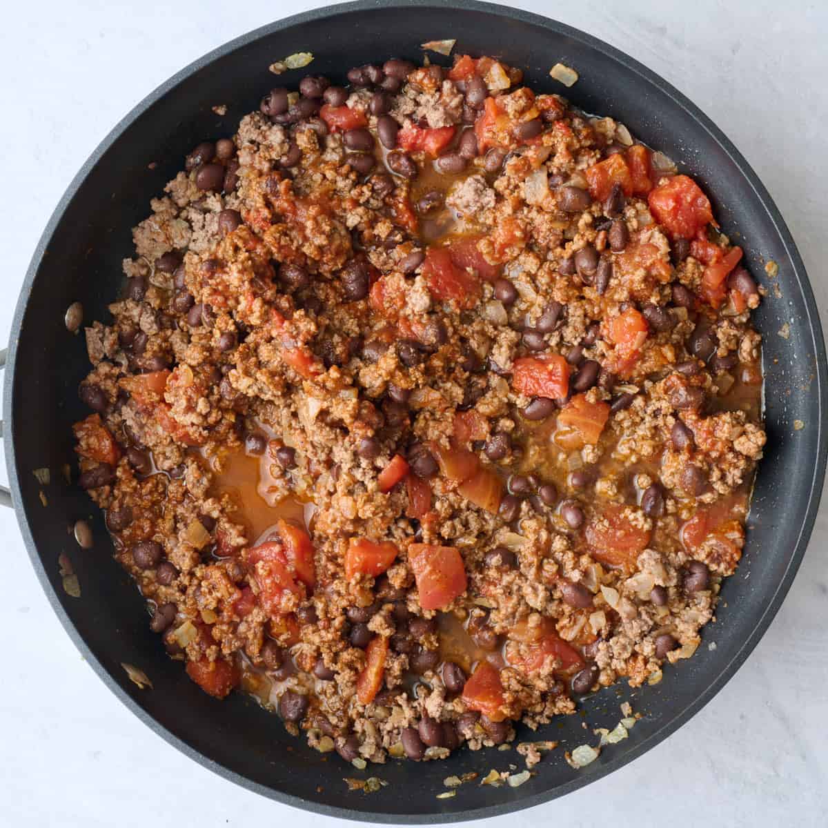Beef in the skillet after cooking with taco seasoning, salt, tomato paste, water, beans, and tomatoes added and combined.