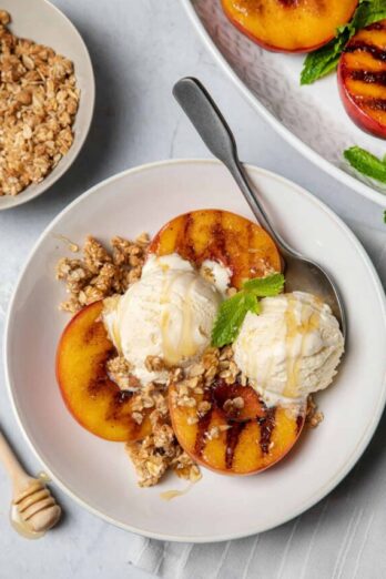 3 grilled peach halves in a bowl topped with ice cream, granola and fresh mint