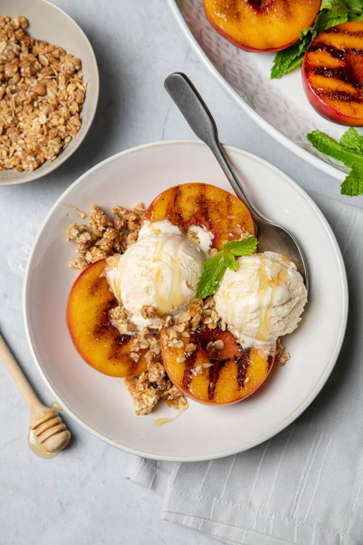 3 grilled peach halves in a bowl topped with ice cream, granola and fresh mint