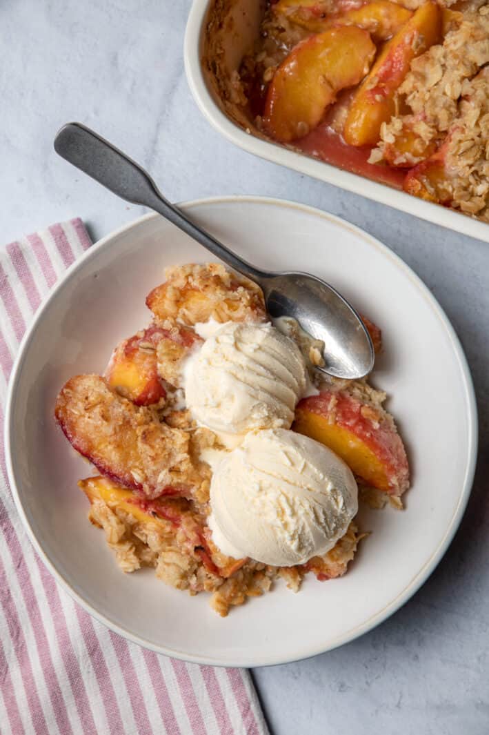 Peach crisp in a bowl served with two scoops of vanilla ice cream.