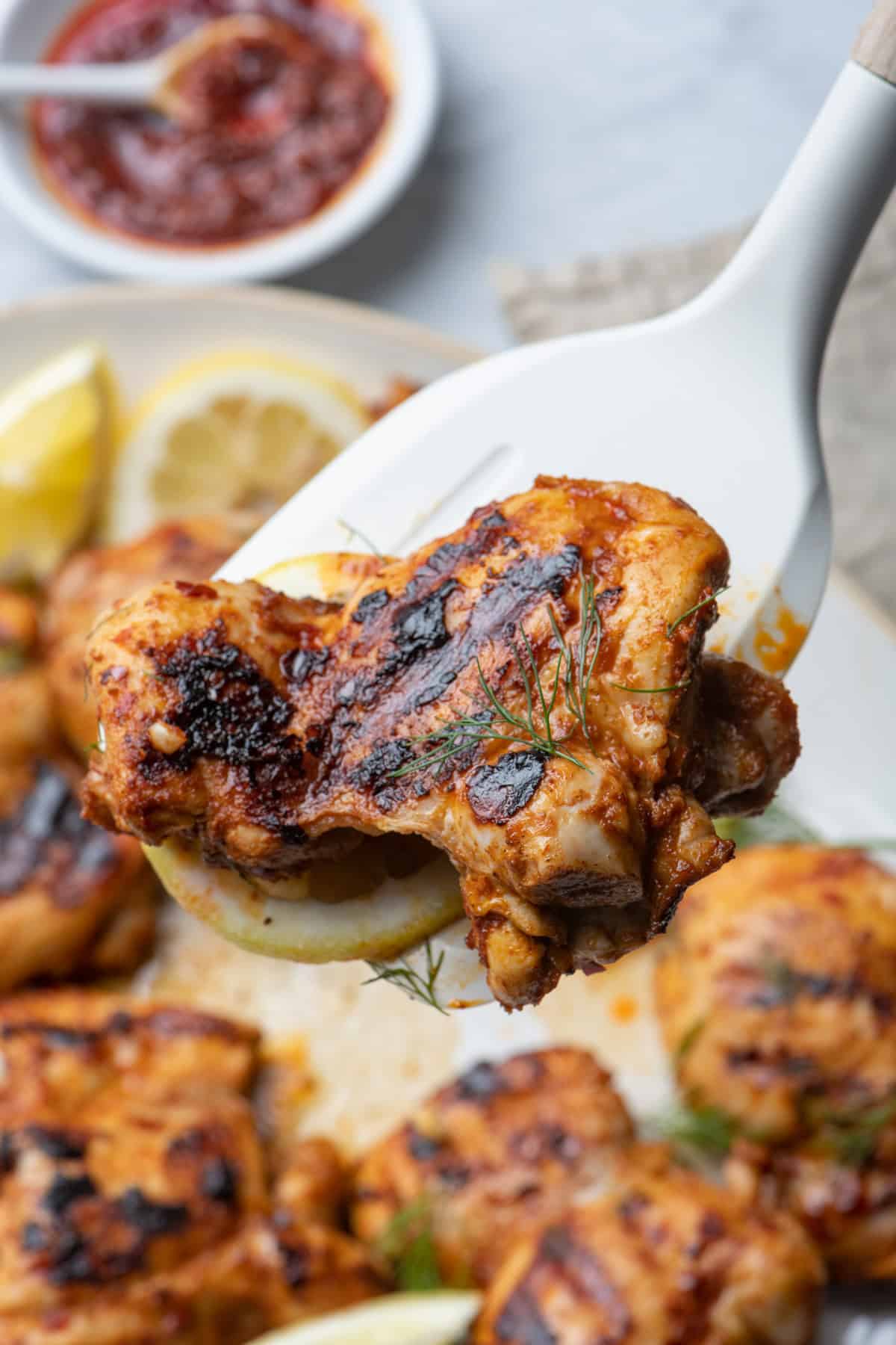 Grilled harissa chicken held up on spatula with more chicken in background.