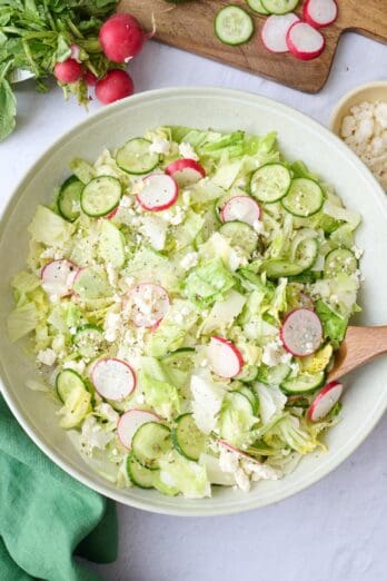 Iceberg salad in a serving bowl garnished with extra feta, serving set dipped inside, and extra ingredients nearby.