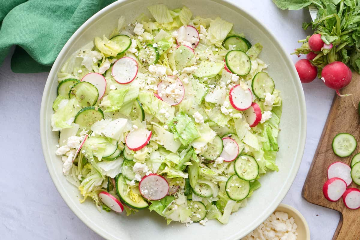 Iceberg salad in a serving bowl garnished with extra feta, serving set dipped inside, and extra ingredients nearby.