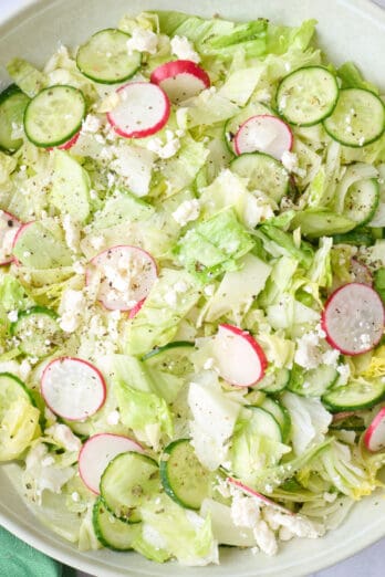 Iceberg salad in a serving bowl garnished with extra feta, serving set dipped inside, and extra ingredients nearby.