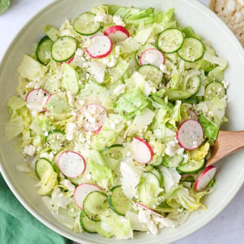 Iceberg salad in a serving bowl garnished with extra feta, serving set dipped inside, and extra ingredients nearby.