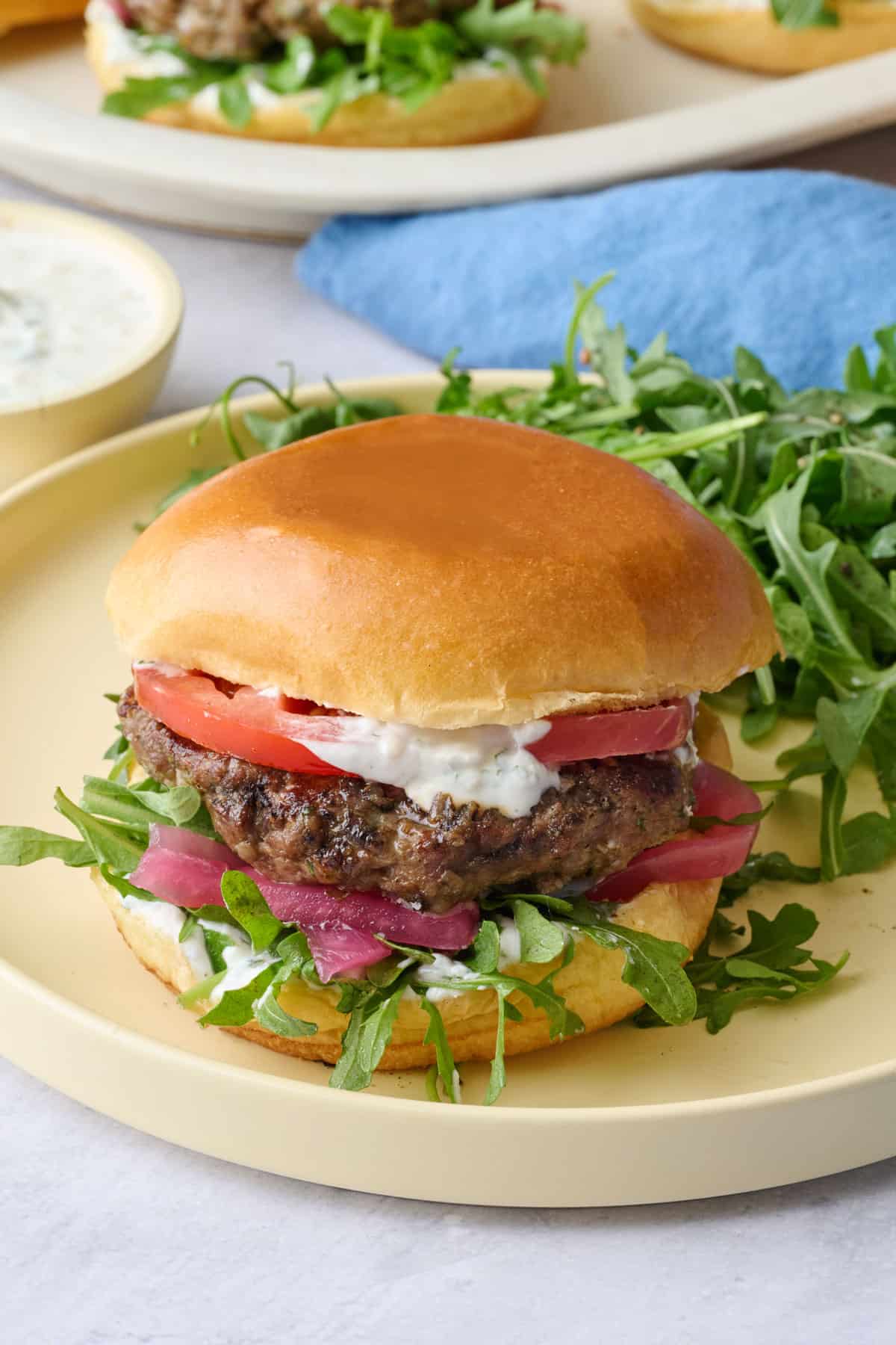 One assembled burger on a plate served with a side of arugula tossed with lemon juice and olive oil and a yogurt dish nearby.