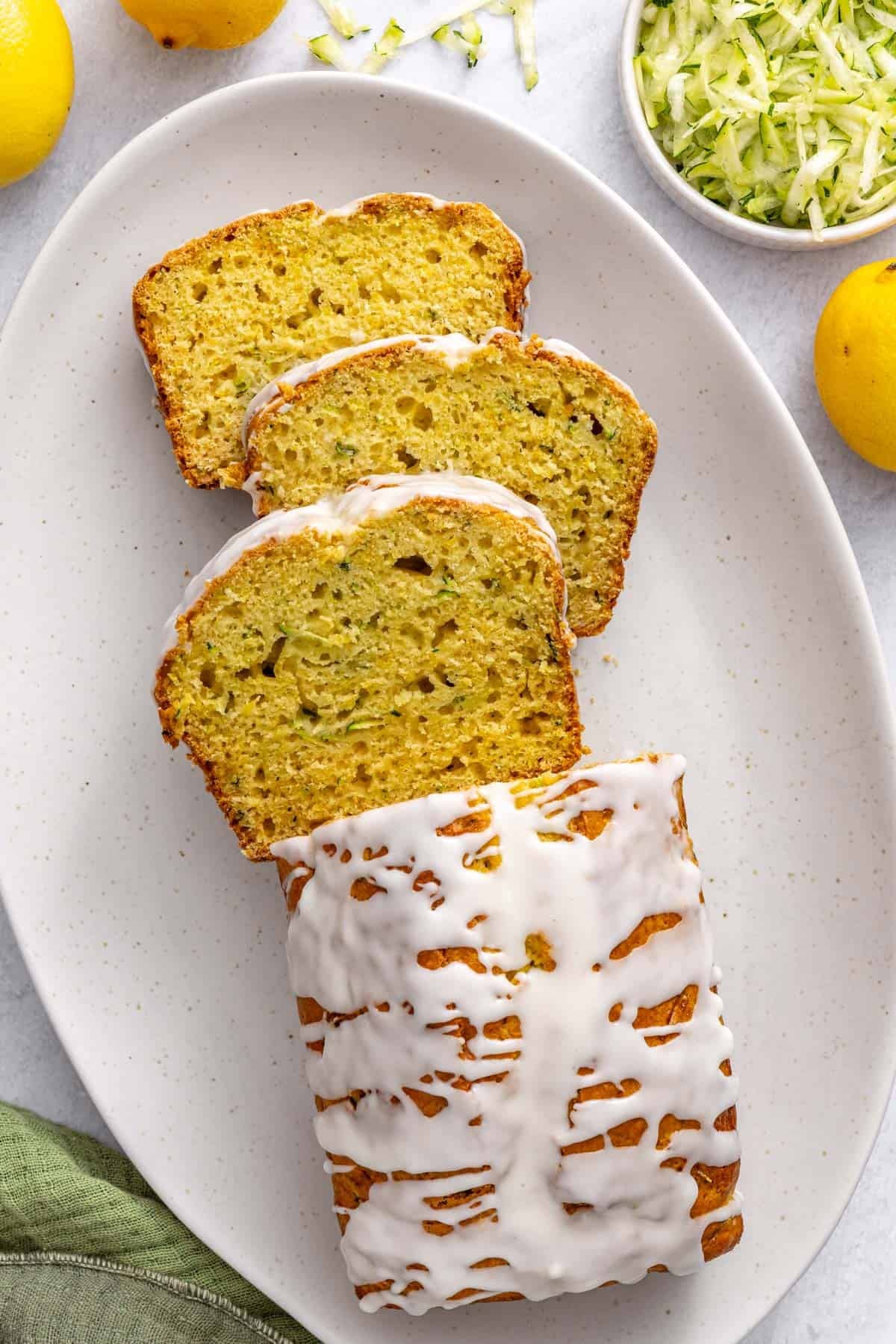 Lemon zucchini loaf on a platter drizzled with icing and garnished lightly with lemon zest. A few thick slices cut and leaning forward.