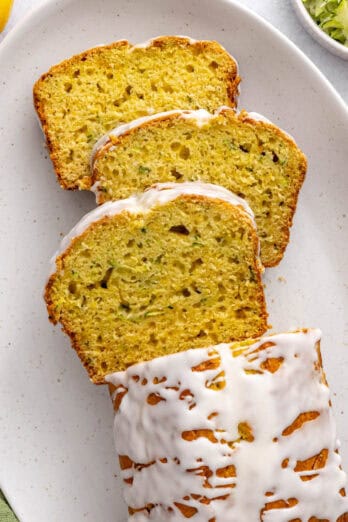 Lemon zucchini loaf on a platter drizzled with icing and garnished lightly with lemon zest. A few thick slices cut and leaning forward.