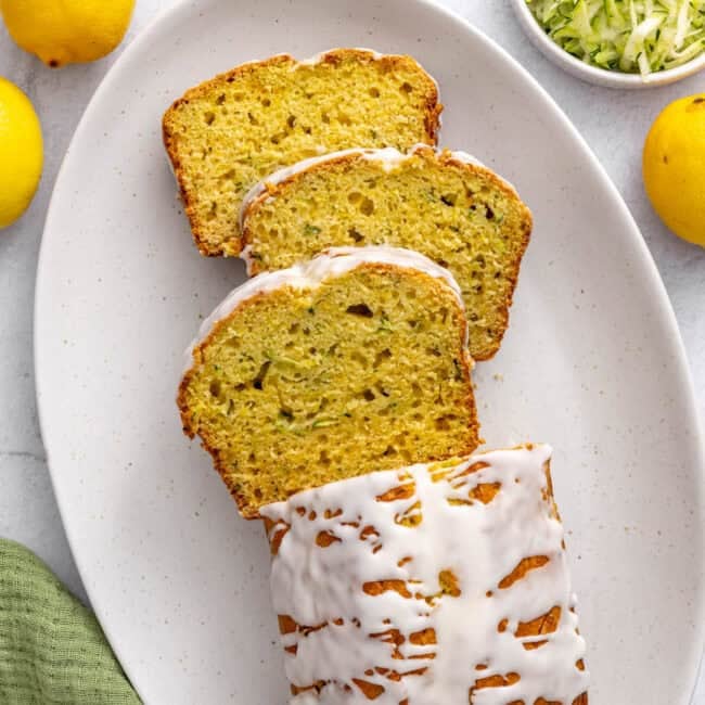 Lemon zucchini loaf on a platter drizzled with icing and garnished lightly with lemon zest. A few thick slices cut and leaning forward.