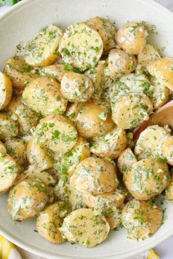 Tahini potato salad in a round serving bowl garnished with extra chopped herbs with lemon wedges in a small dish nearby, serving spoon nearby.