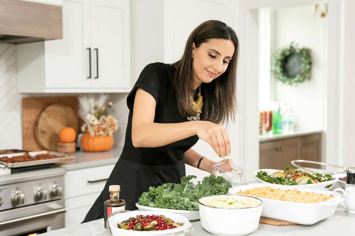 Yumna standing over side dishes sprinkling on the final touches of fresh herbs to a bowl of mashed potatoes.