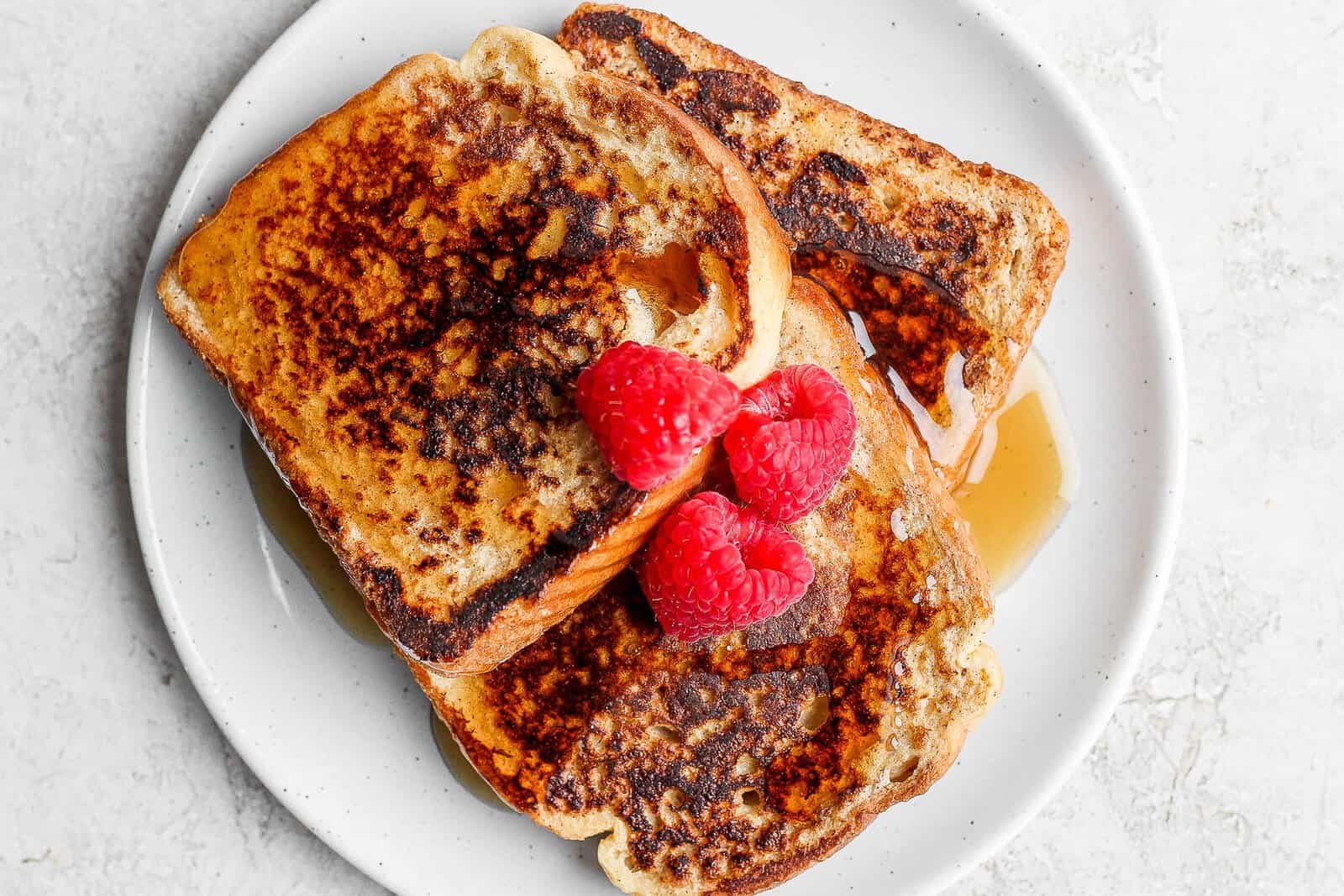 Three pieces of french toast on a plate topped with raspberries and syrup.
