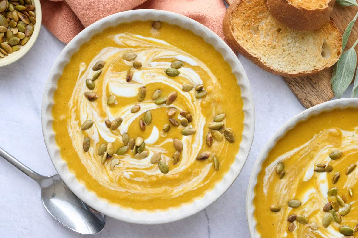 Two bowls of pumpkin soup garnished with an extra swirl of yogurt and pumpkin seeds.