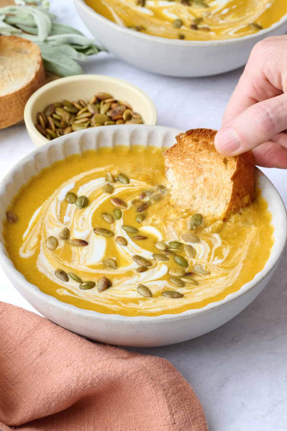 Two bowls of pumpkin soup garnished with an extra swirl of yogurt and pumpkin seeds and a hand dipping a piece of bread into one bowl.