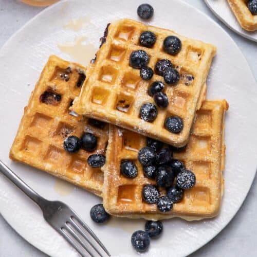 Plate of waffles with blueberries, powdered sugar and maple syrup on top.